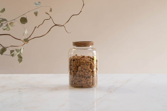 A large glass food storage jar filled with cereal sits on a marble surface with a dried twig in the foreground.
