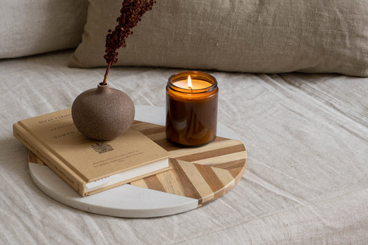 A lit candle sits on top of a serving board, along with a book and small vase with flower.