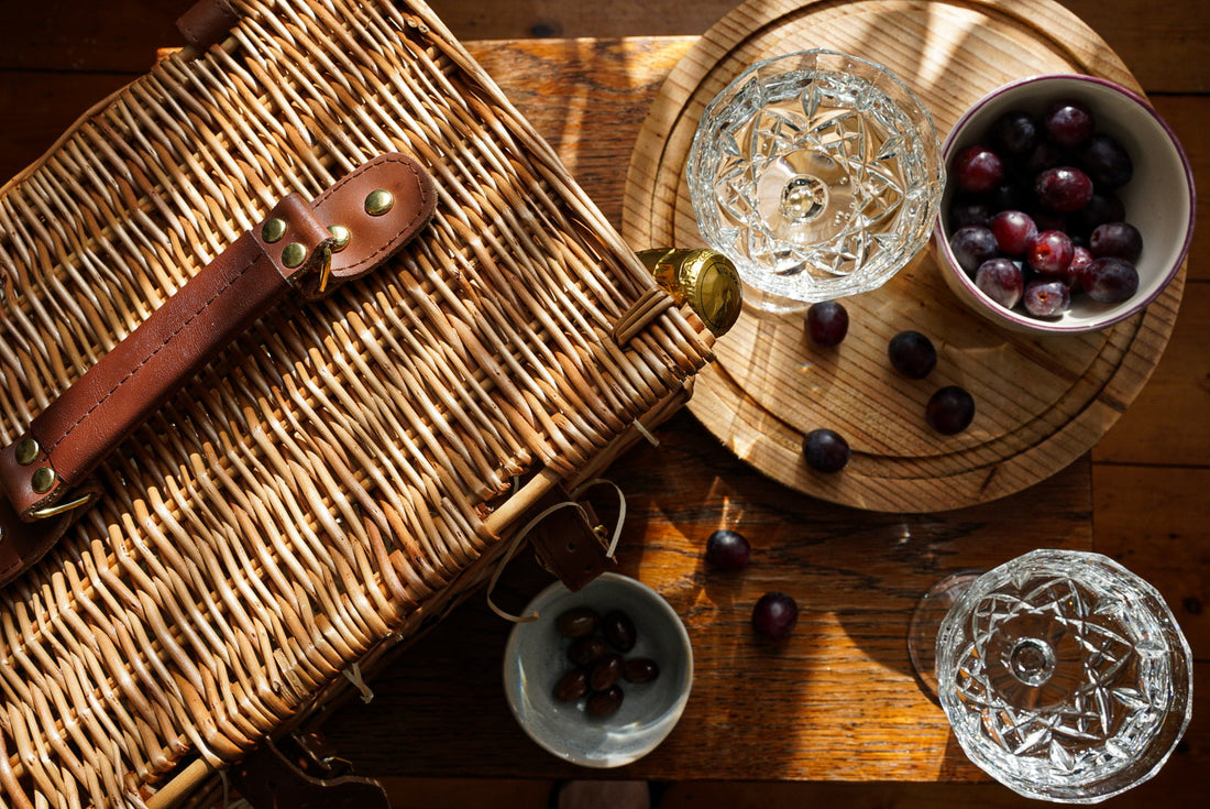 Ariel view on a picnic table including a basket with a bottle of champagne poking out, champagne saucers, a wooden serving board and some grapes in a bowl